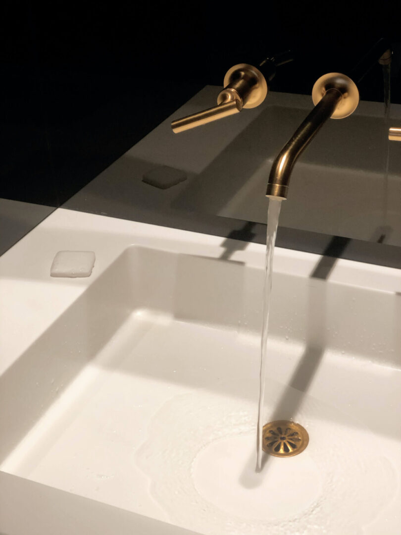 A white sink with gold faucets and a black wall.